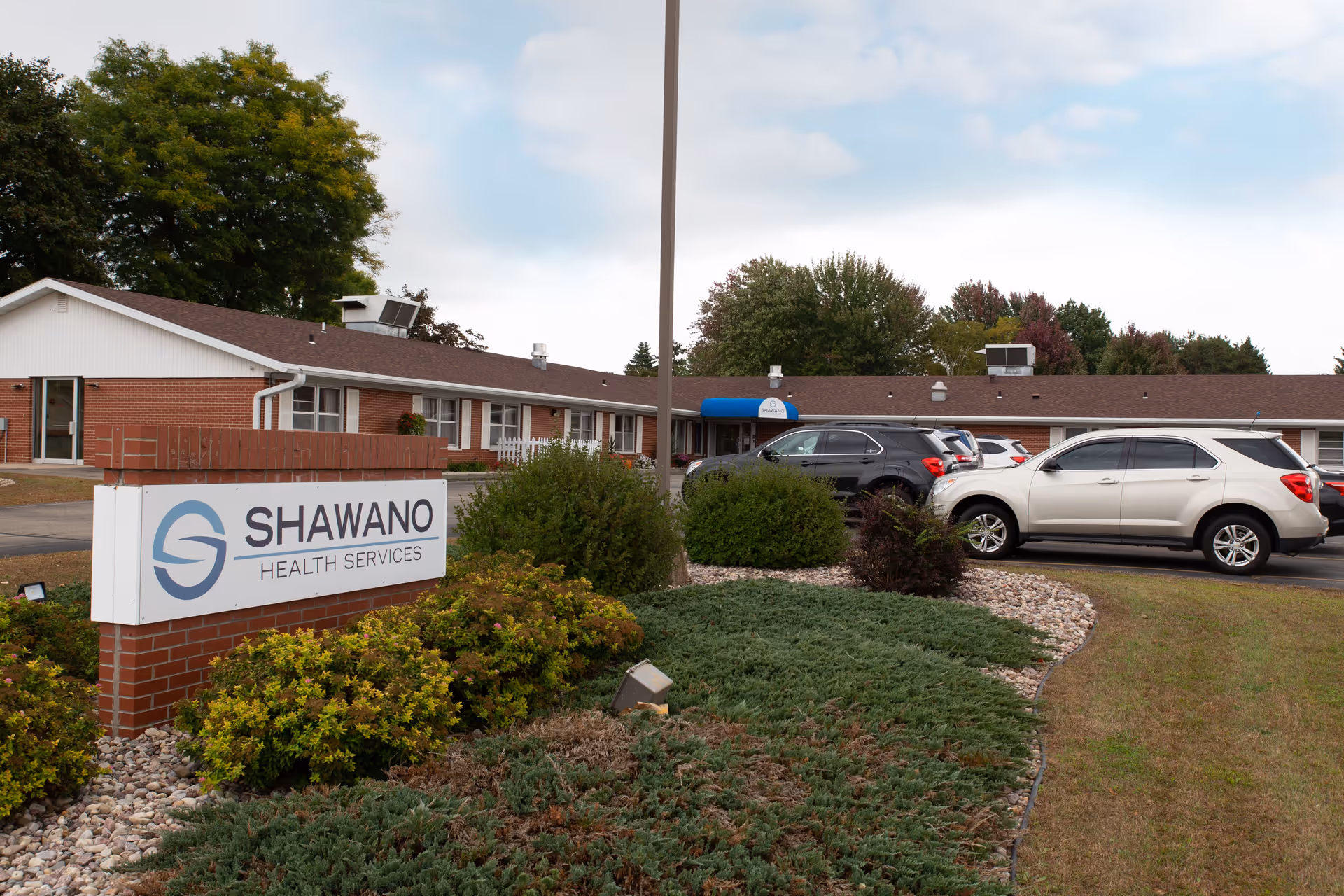 Exterior view of Shawano Health Services building with a brick sign in the foreground and several parked cars near the entrance. The building is single-story with a brown roof and white siding, surrounded by greenery and trees under a partly cloudy sky.