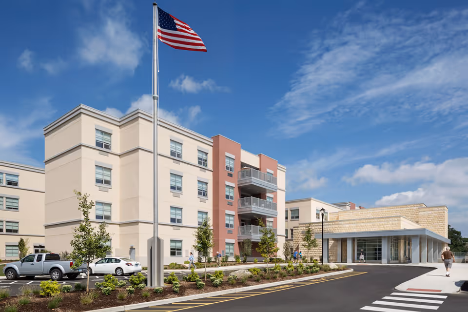 Modern senior living building with flagpole, parking, and landscaped entrance under a bright blue sky.