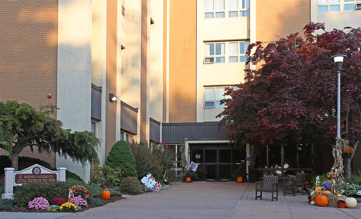 Entrance to St. Joseph's Residence at Little Sisters of the Poor, showing a multi-story brick building with a pathway leading to glass doors. The area is decorated with pumpkins, flowers, and autumn-themed decorations, with trees and benches nearby.