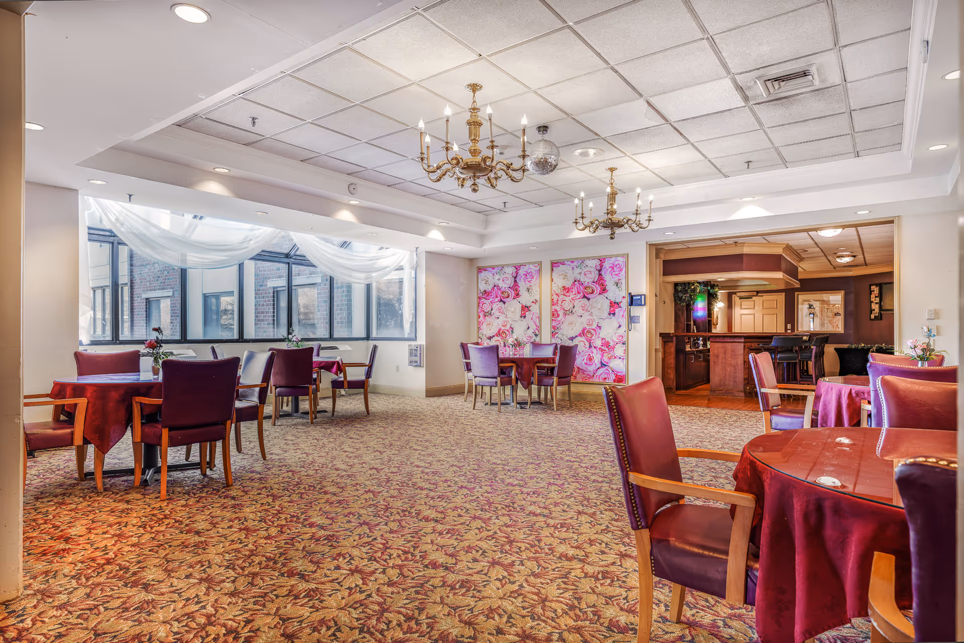 A spacious senior living dining area with burgundy tablecloths, purple chairs, chandeliers, and windows.