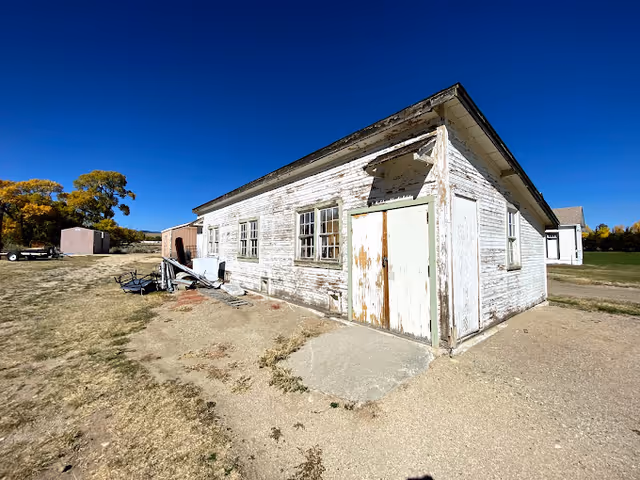 Weathered white wooden single-story building with peeling paint and a greenish door on a grassy lot under a clear blue sky.
