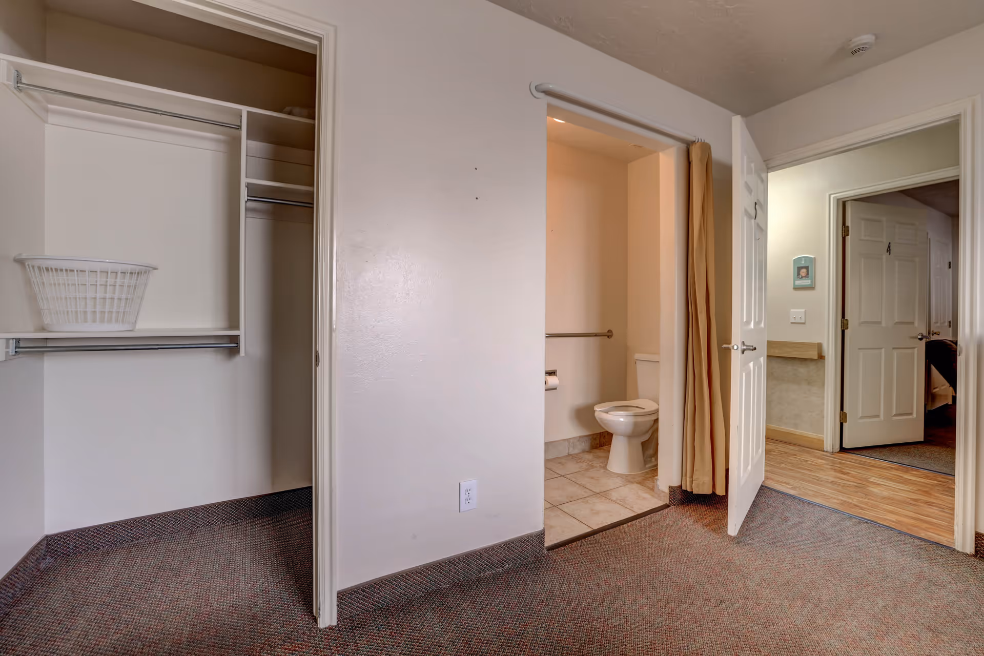 Interior view of a senior living facility room showing an open closet with a white laundry basket on the shelf, an open bathroom door revealing a toilet and grab bar, and a hallway with doors leading to other rooms.