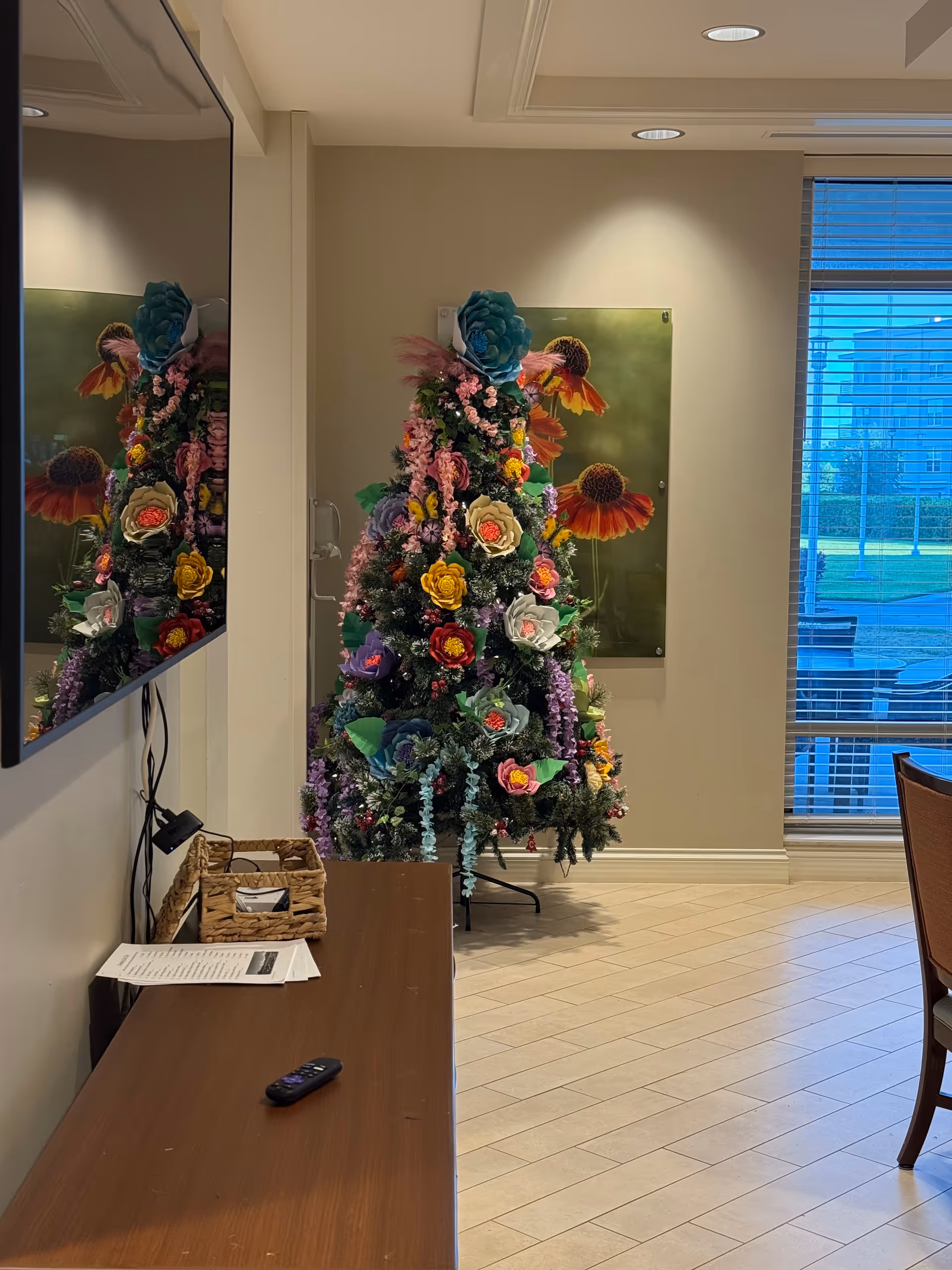 Indoor lobby with a colorful flower-adorned Christmas tree near a window.