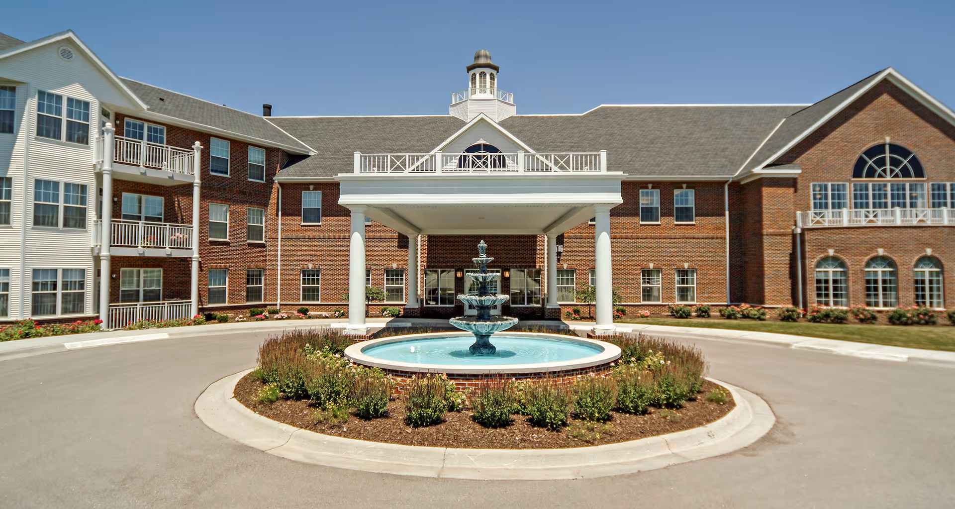 Front entrance of a brick senior living building with a covered porte-cochère and a circular fountain in the driveway.