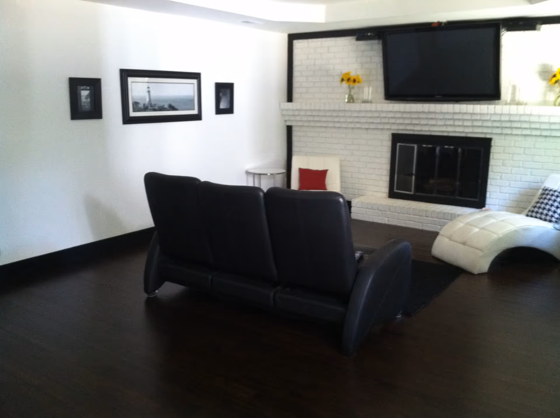 A living room with a black leather couch facing a white brick fireplace. Above the fireplace is a mounted flat-screen TV. There are two vases with yellow flowers on the mantel. To the right of the fireplace is a white chaise lounge with a black and white pillow, and a white chair with a red pillow. The floor is dark wood, and there are three framed pictures on the white wall to the left.
