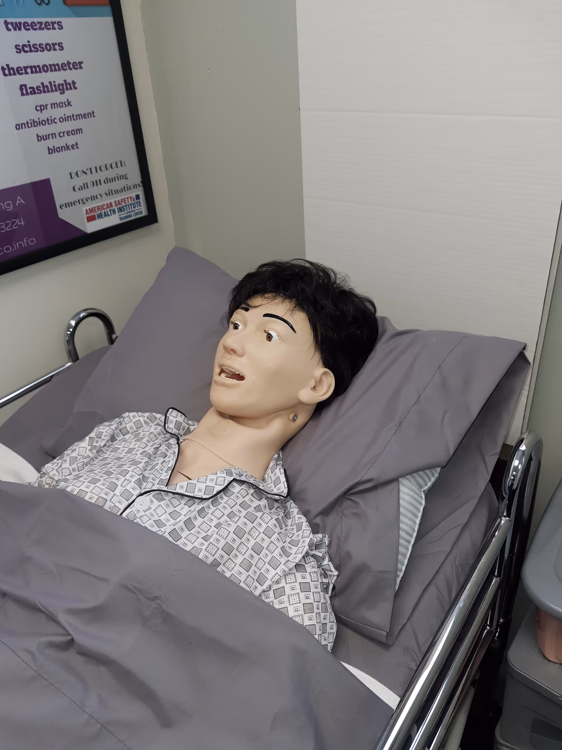Medical training mannequin lying in a hospital-style bed inside a clinical room.