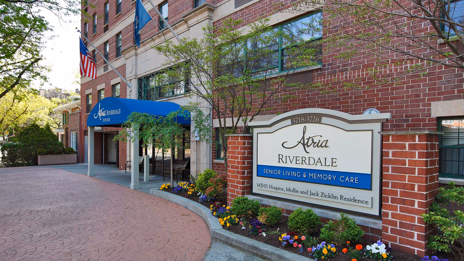 Exterior view of Atria Riverdale senior living and memory care facility with a brick building, blue awning over the entrance, American flag, and a landscaped area with flowers and shrubs near the sign.