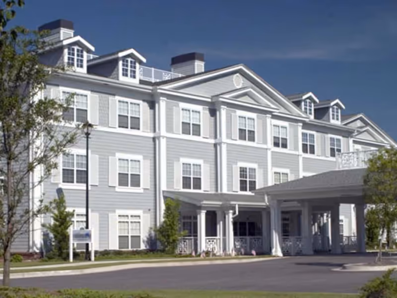 Front exterior of a three-story light-gray senior living building with white trim, multiple windows, dormers, and a covered porte-cochère entrance.