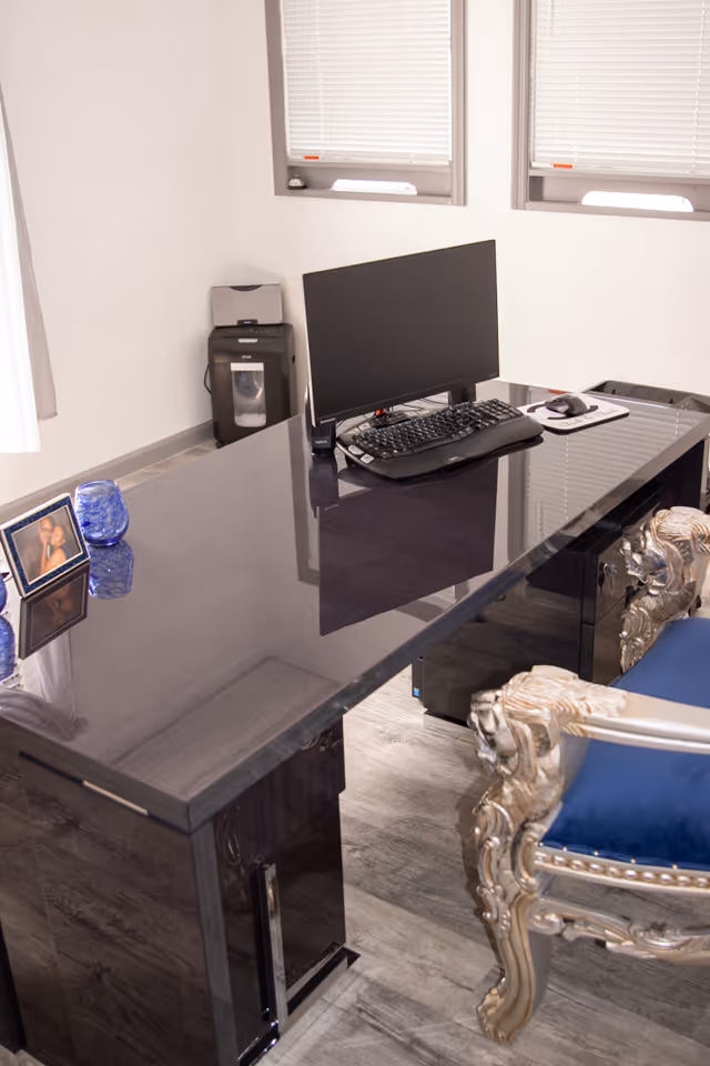 Glass-topped desk with computer setup, ornate silver chair, and windows.