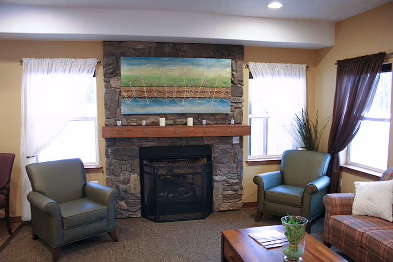 A cozy living room with a stone fireplace in the center, flanked by two windows with white curtains. Two green armchairs are positioned on either side of the fireplace, and a plaid sofa with a white pillow is on the right. A wooden coffee table with a glass vase and some magazines is in front of the sofa. Above the fireplace is a colorful painting with abstract designs.