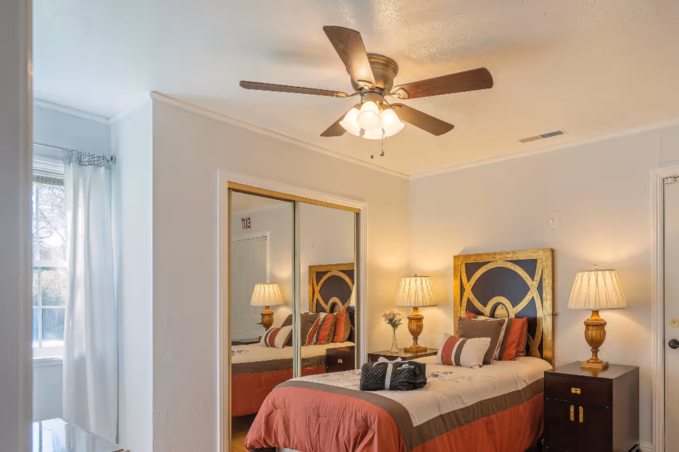Bright single bedroom with a decorative headboard, two bedside lamps, mirrored closet doors, a ceiling fan, and a window with curtains.