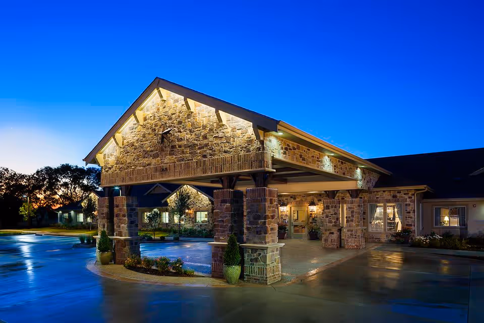 Exterior view of Arbor Hills Memory Care Community at dusk, showing a stone and brick entrance with a covered driveway and warm lighting illuminating the building and surrounding landscaping.
