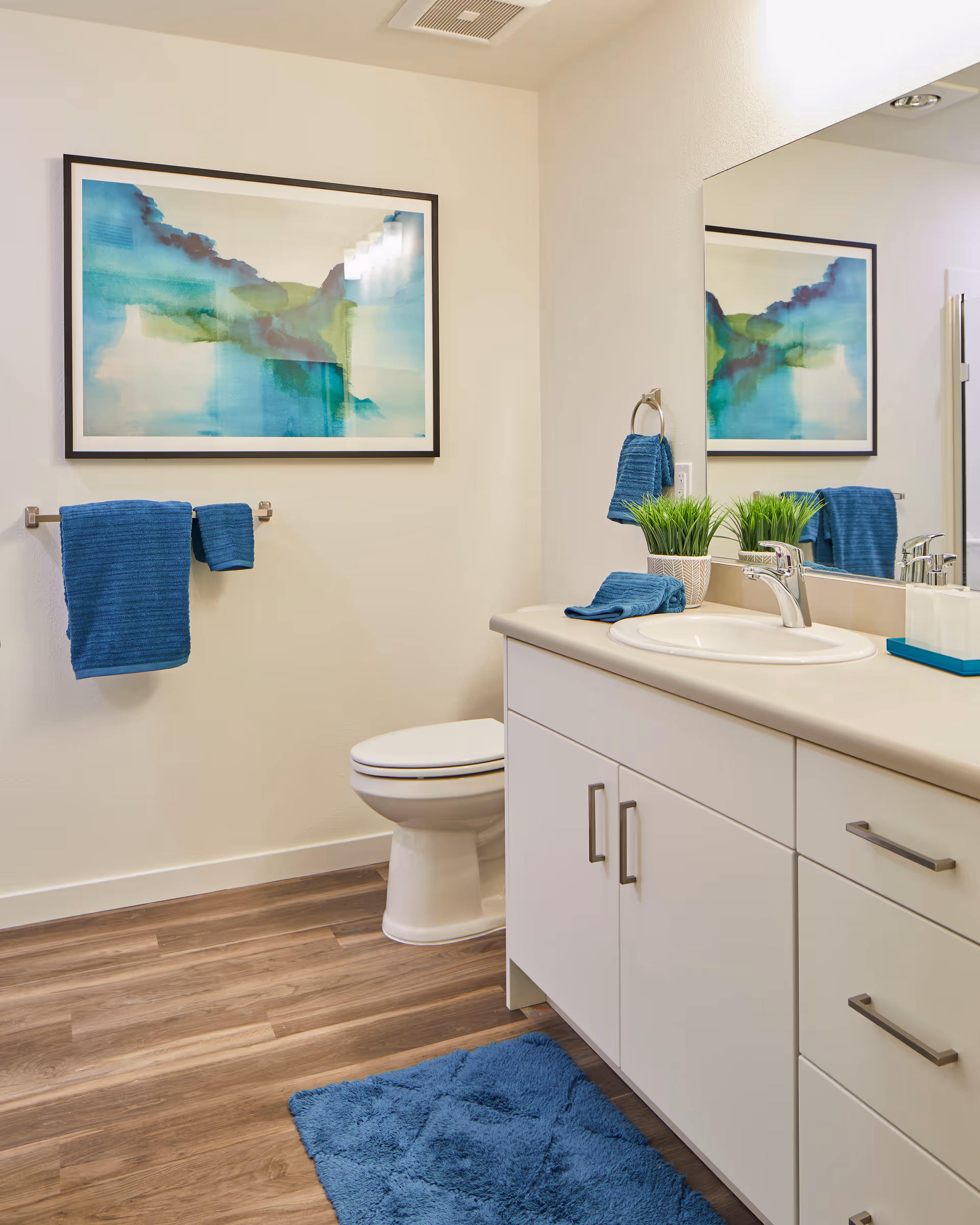 A clean bathroom with a white toilet, white vanity with a sink, and a large mirror. Blue towels hang on a towel rack and a ring, with a blue bath mat on the wooden floor. A framed abstract watercolor painting in shades of blue and green is on the wall.