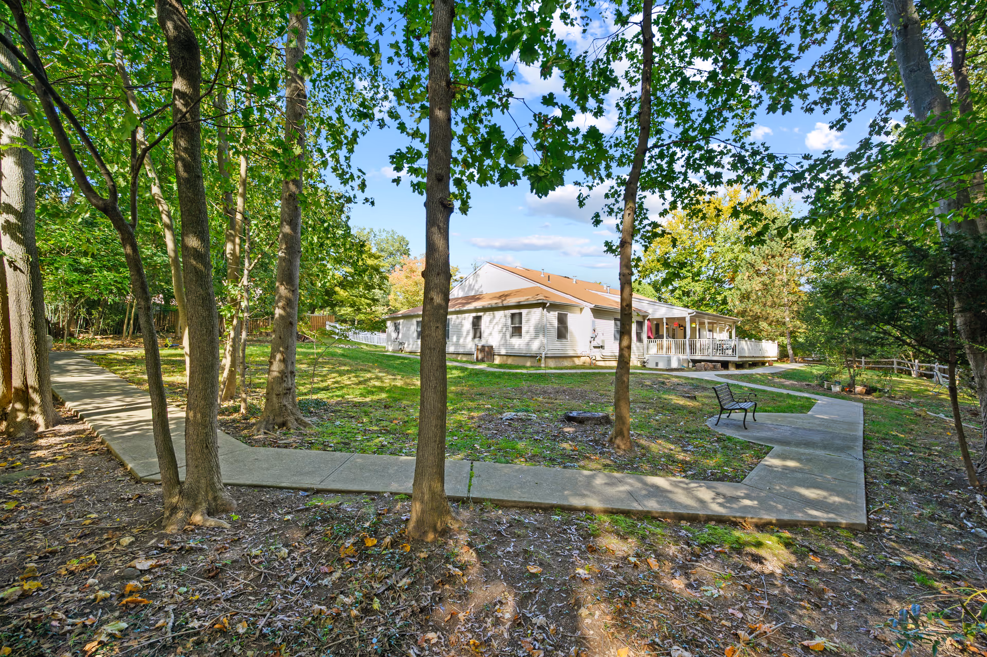 Single-story memory care building set in a wooded yard with concrete walkways and a bench.