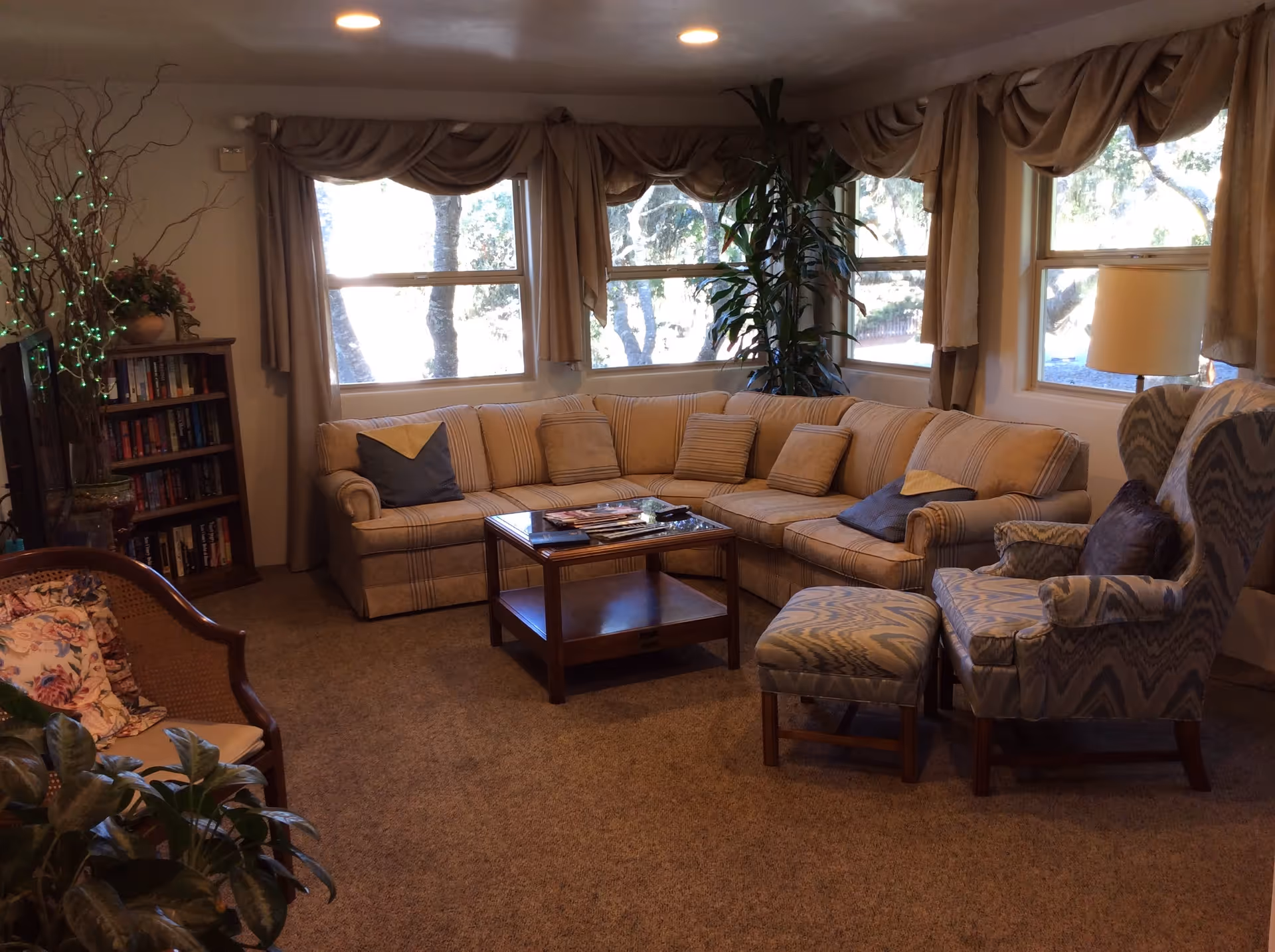 A cozy living room with a large beige sectional sofa adorned with cushions, a wooden coffee table with magazines on top, a patterned armchair with a matching ottoman, a wooden bookshelf filled with books, several plants, and large windows with beige curtains letting in natural light.