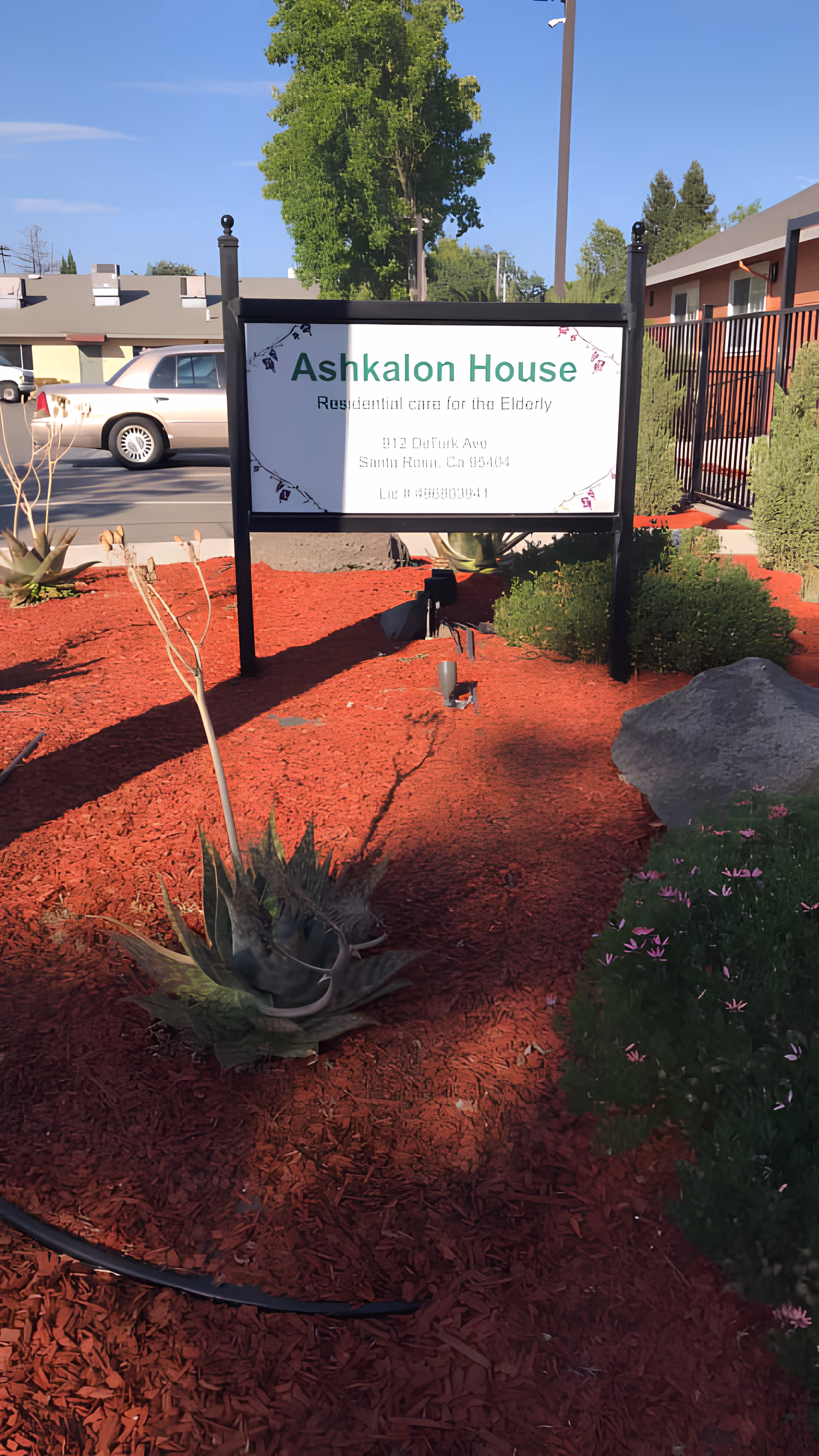 An outdoor sign reading "Ashkalon House" stands in a landscaped front yard with red mulch, succulents, and the facility building in the background.