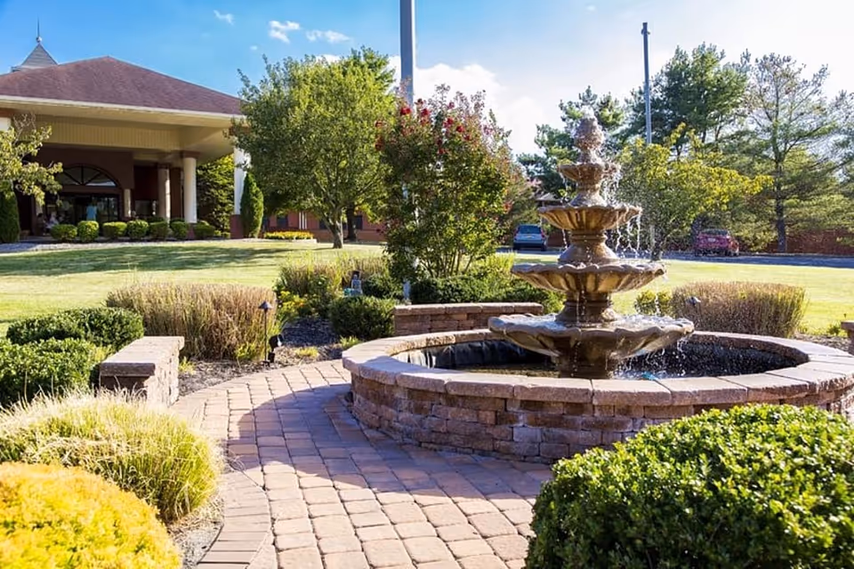 Stone fountain centerpiece with brick walkway and landscaped shrubs in front of a senior facility.
