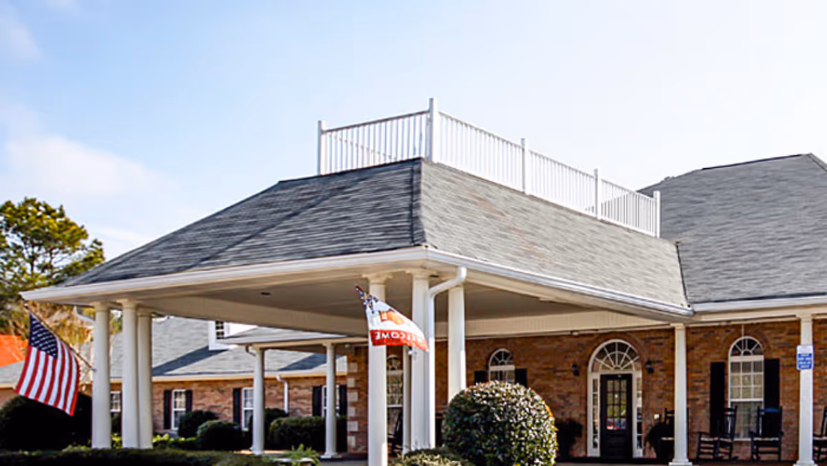 Exterior view of a senior living facility building with a covered entrance supported by white columns, brick walls, and multiple windows. There are two flags displayed near the entrance, including an American flag. The sky is clear and blue.