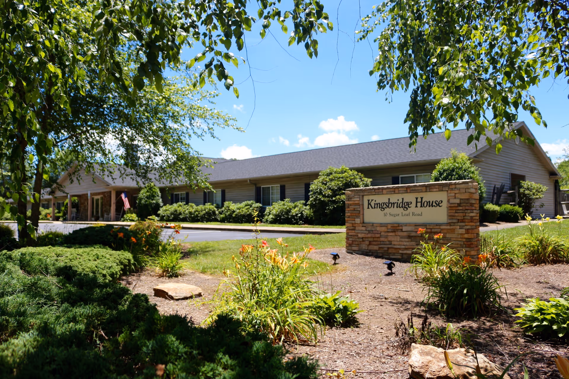 Exterior view of Kingsbridge House, a single-story building with a dark roof and beige siding, surrounded by green bushes and trees under a blue sky with some clouds. In the foreground, there is a stone sign with the name 'Kingsbridge House' and the address '10 Sugar Loaf Road' surrounded by landscaped plants and flowers.
