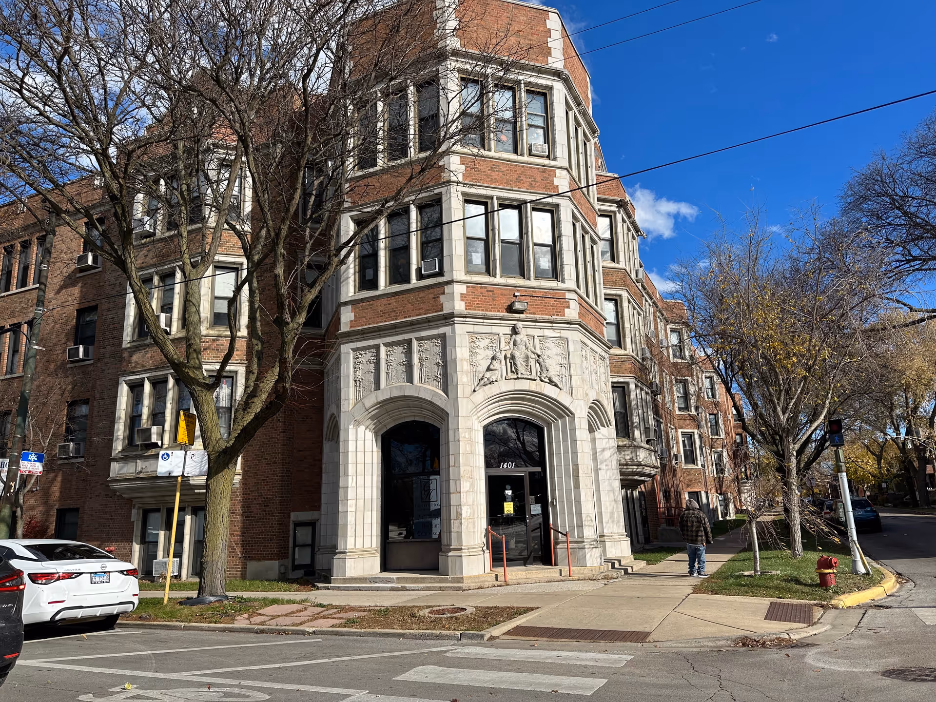Exterior view of a multi-story brick building with decorative stonework around the entrance. The building is located on a street corner with a sidewalk, trees without leaves, parked cars, and a person walking on the sidewalk. The sky is partly cloudy with blue patches.