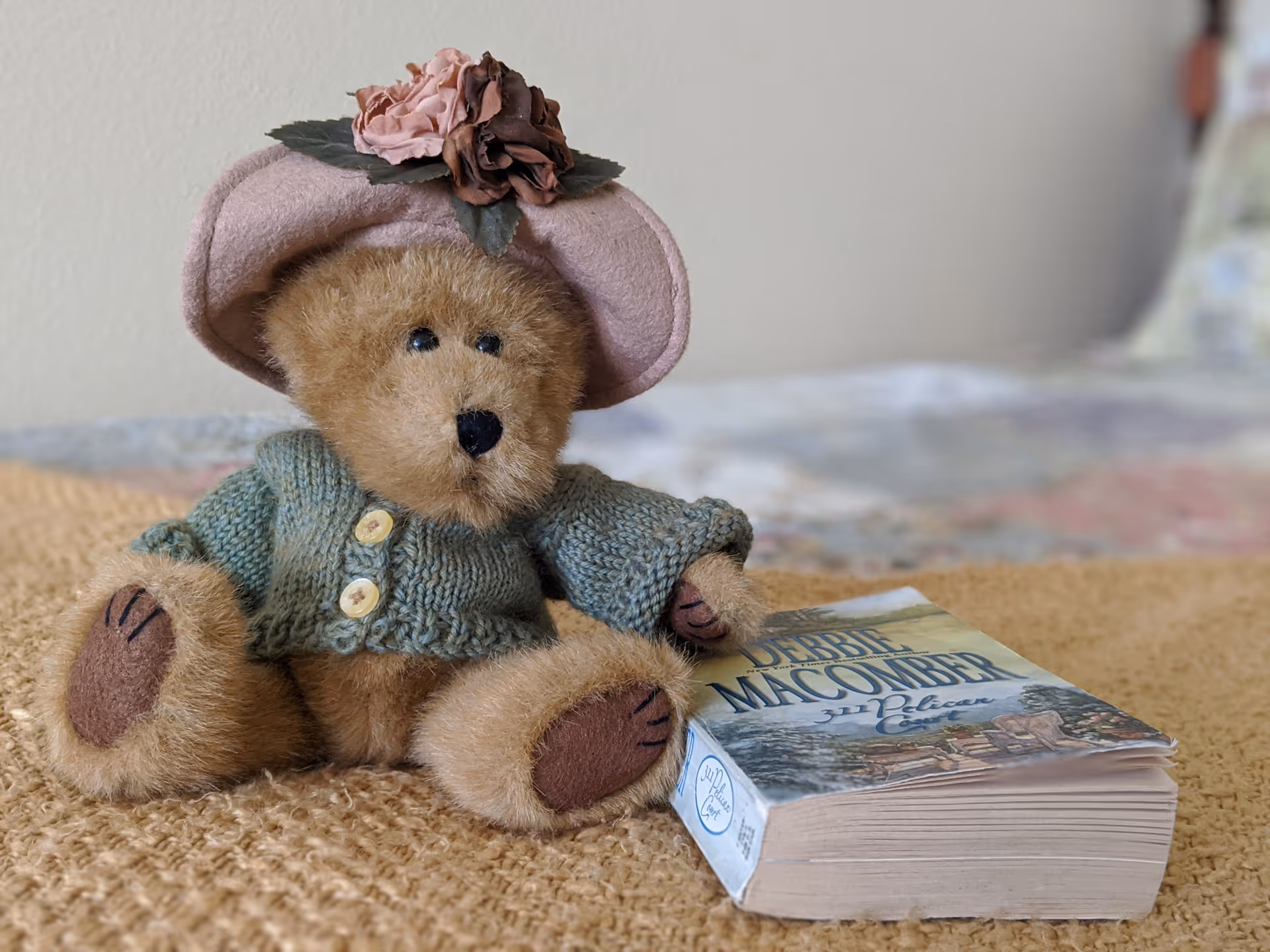 A small teddy bear wearing a pink hat with flowers and a green knitted sweater sitting on a textured beige blanket next to a paperback book titled 'Debbie Macomber 32 Pelican Court'. The background shows a blurred bed with a quilted bedspread.