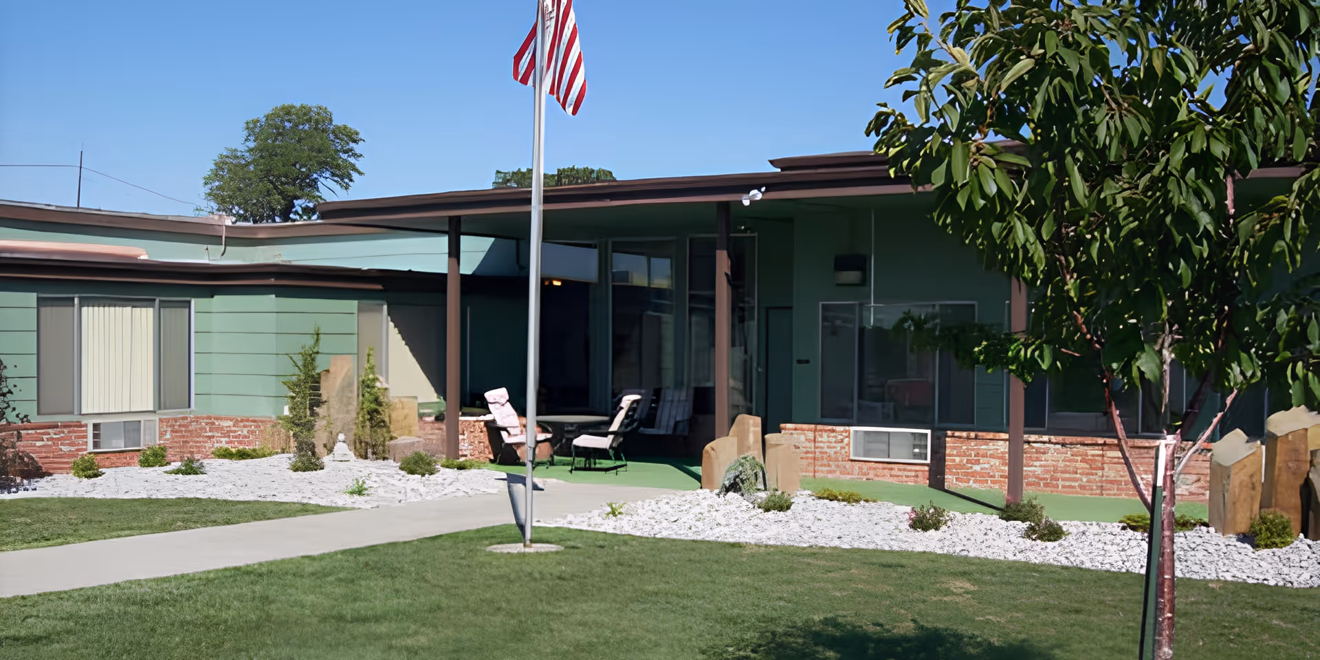 Exterior courtyard and front of a single-story senior living building with an American flag, lawn, rock landscaping, and patio chairs.