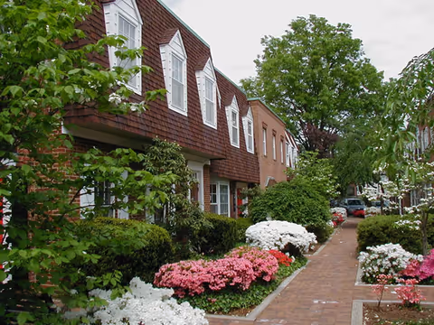 Brick walkway runs beside a landscaped exterior of a senior living building.