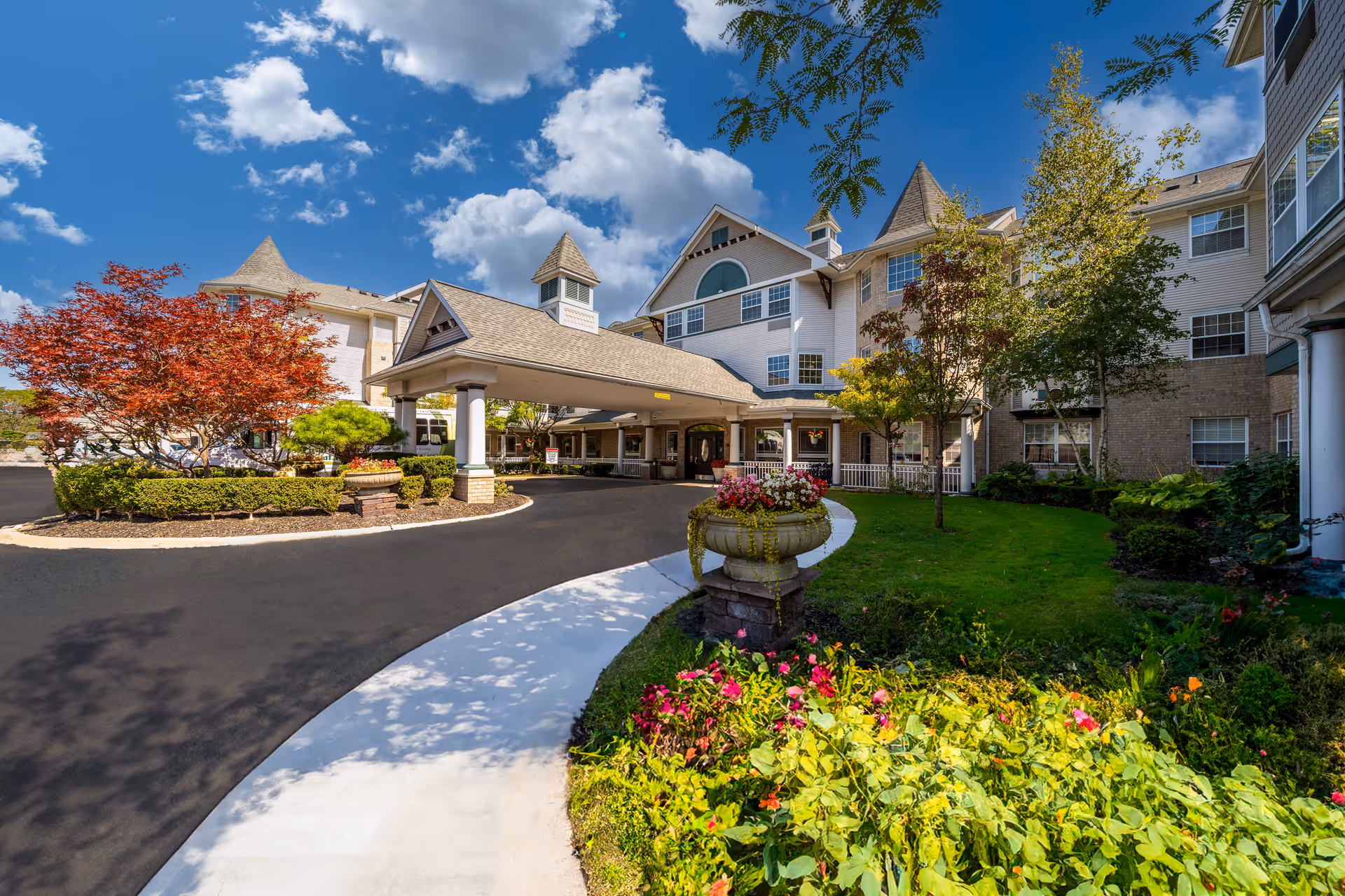 Front entrance of a senior living building with a covered porte-cochere, landscaped driveway and colorful flower beds under a blue sky.