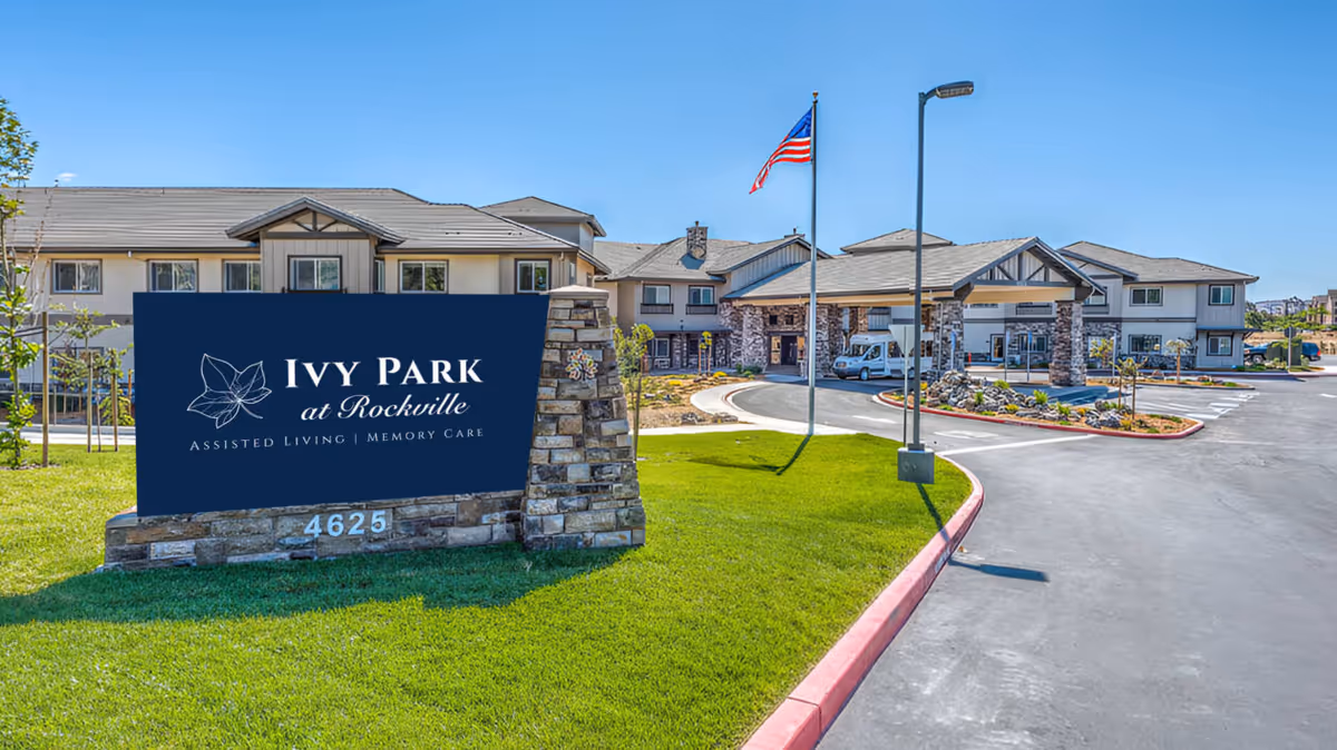 Exterior view of Ivy Park at Rockville assisted living and memory care facility, showing the main building, a driveway with a circular entrance, landscaped green lawn, and an American flag on a flagpole.