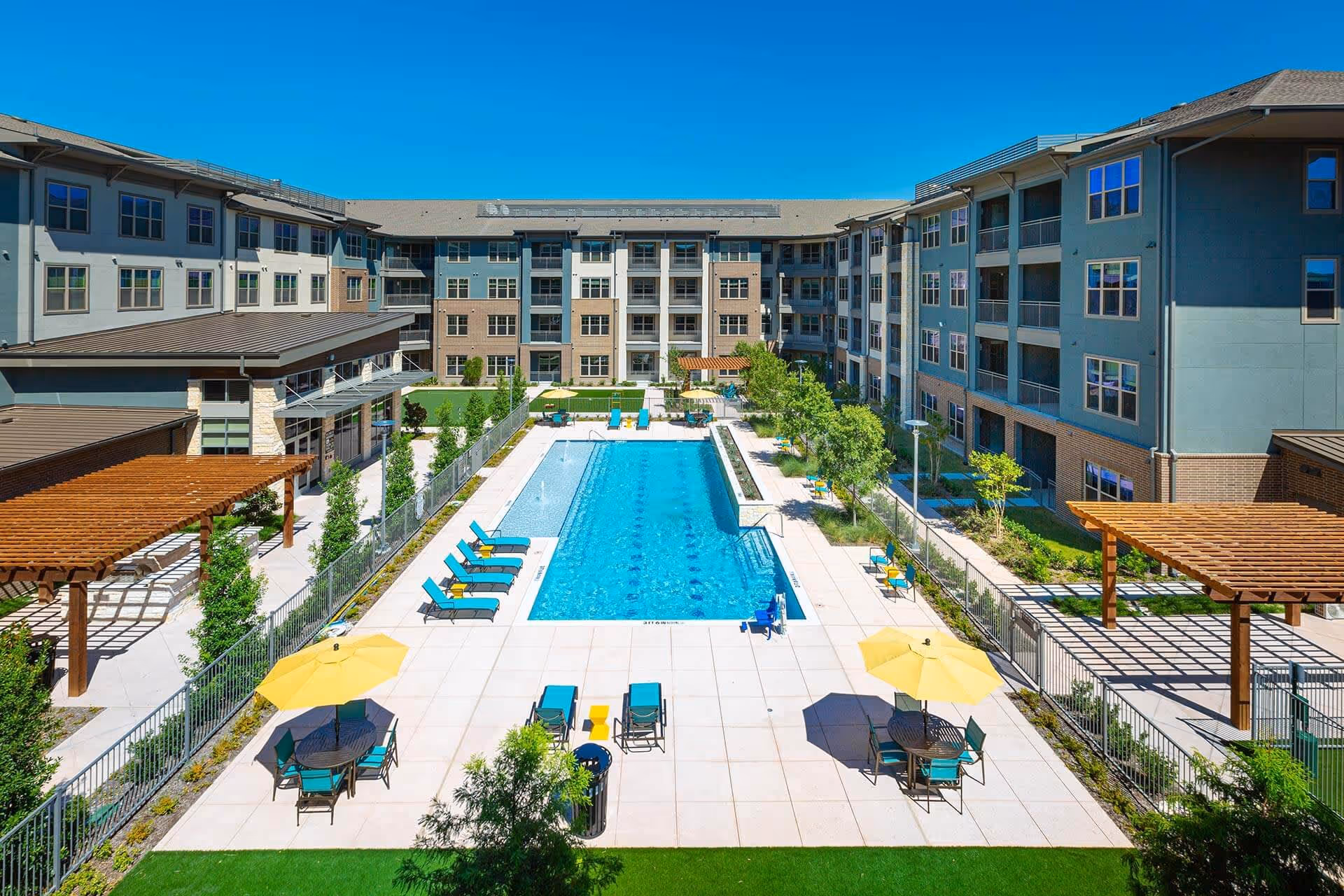 Outdoor swimming pool area at The Orchards Market Plaza with lounge chairs, tables with yellow umbrellas, pergolas, and a surrounding four-story residential building under a clear blue sky.