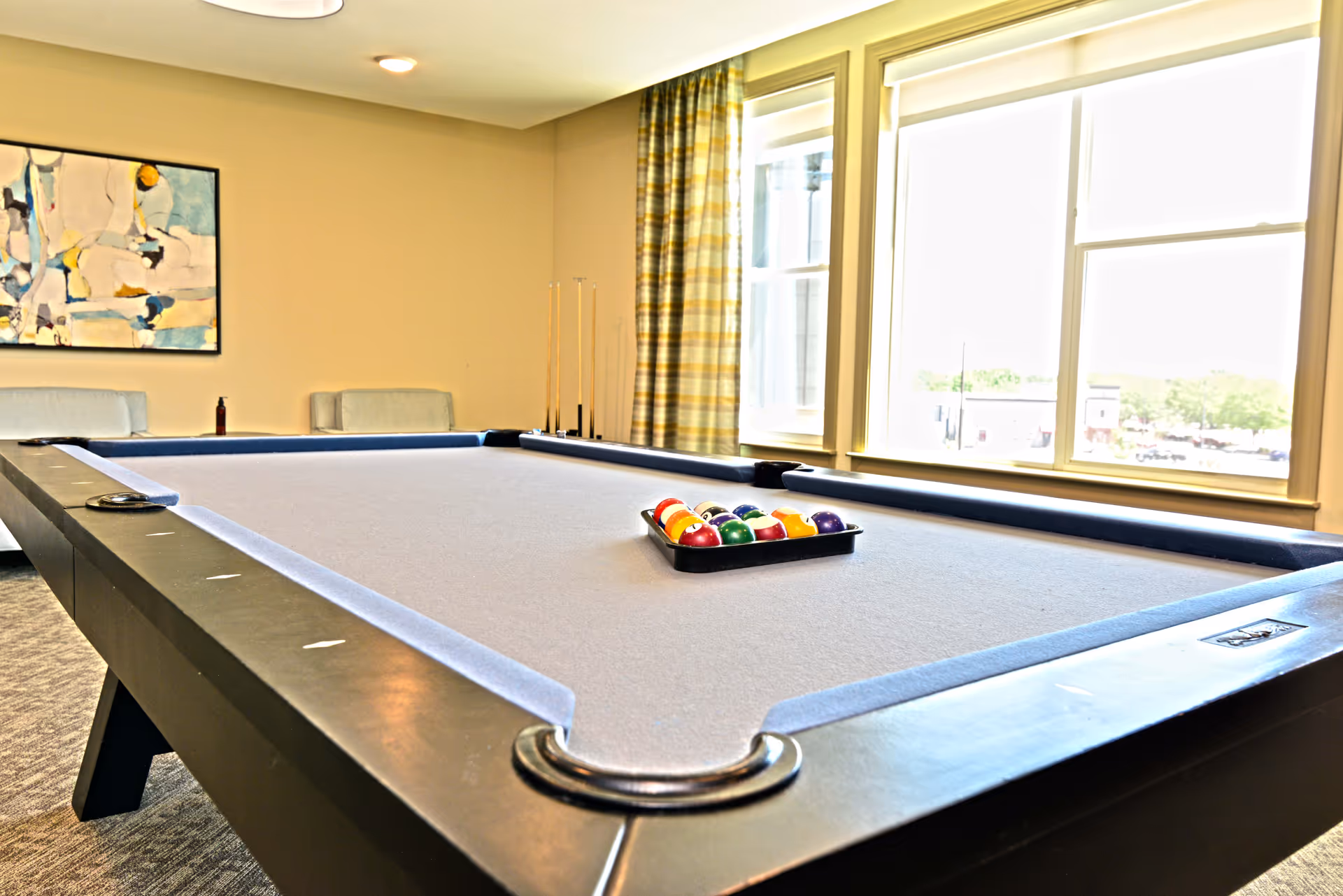 Bright recreation room featuring a pool table, balls on a tray, windows, and an abstract painting.
