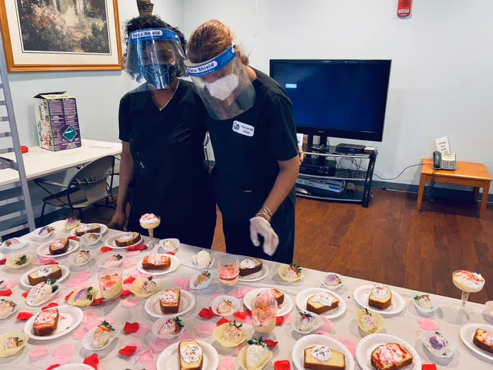 Two staff members wearing black scrubs, face masks, and clear face shields stand behind a table filled with plates of decorated desserts in a room with wooden flooring, a TV on a stand, and a small table with a phone.