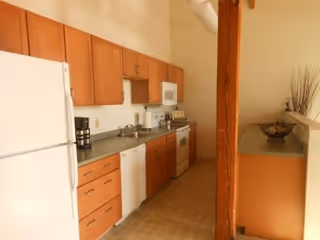 A kitchen with wooden cabinets, a white refrigerator, dishwasher, microwave, and stove. The countertops are gray, and there is a coffee maker on the counter. The floor is tiled, and a wooden beam is visible near the counter with a decorative bowl and tall dried plants.