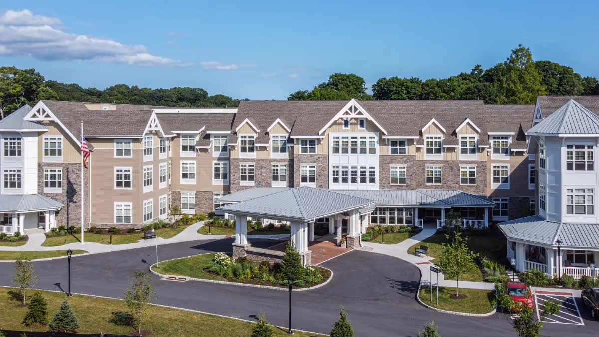 Front exterior of a multi-story senior living building with a covered entrance, American flag, and landscaped driveway.