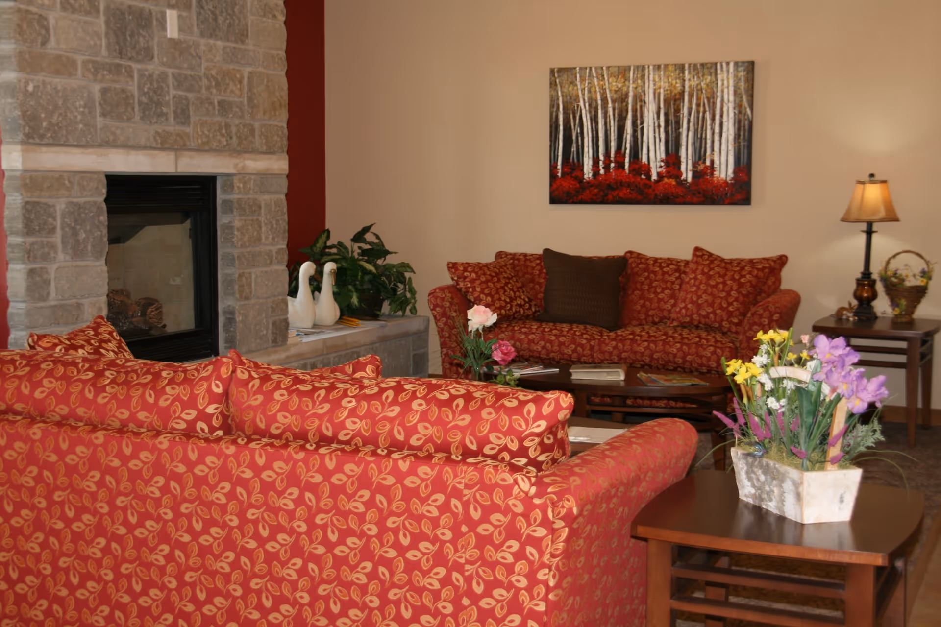 Comfortable living room with matching red patterned sofas, a stone fireplace, coffee and side tables with flowers and a lamp, and a forest painting on the wall.