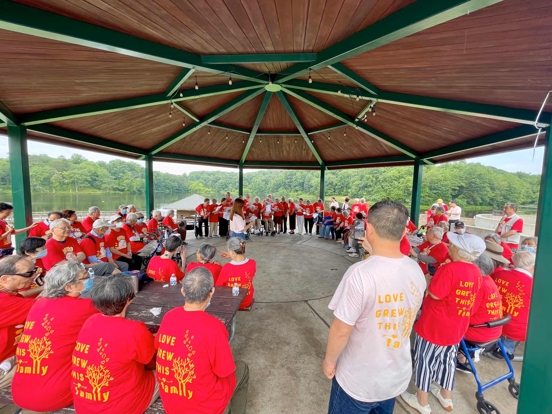 Seniors in red shirts form a circle under a green gazebo by a lake.