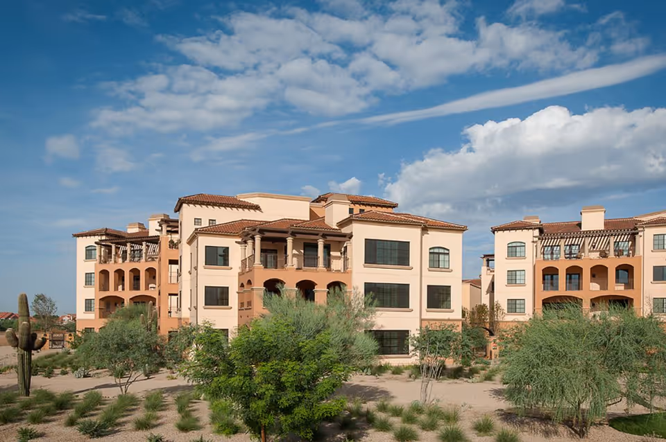 Mediterranean-style multi-story residential building with balconies set in desert landscaping under a partly cloudy sky.