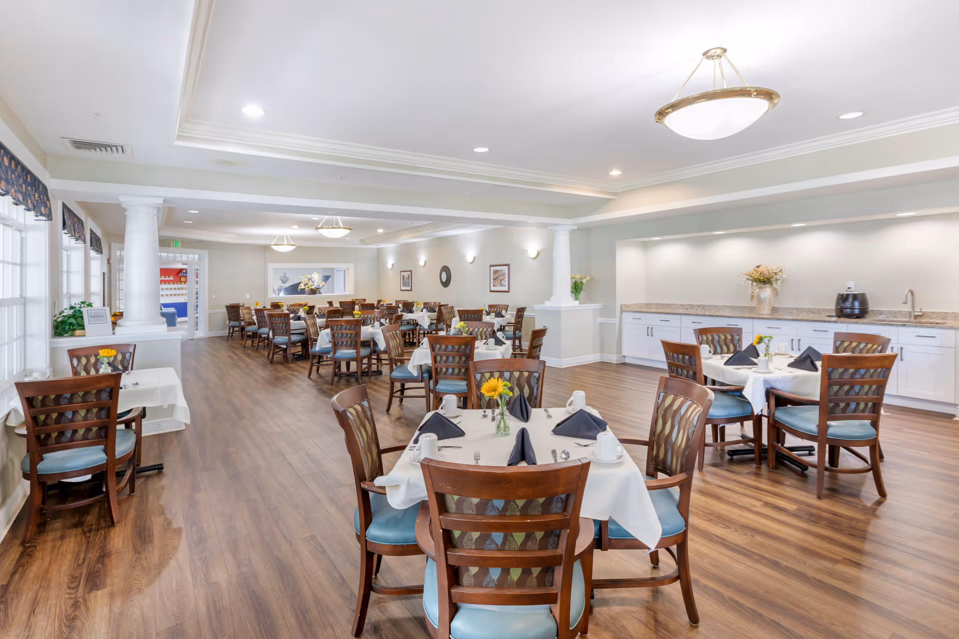 A spacious dining room with multiple wooden tables and chairs arranged neatly. Each table is covered with a white tablecloth and set with black napkins, white cups, and silverware. The room has wooden flooring, white walls, and large windows on the left side letting in natural light. There are decorative columns and ceiling lights, along with a countertop and cabinets on the right side with a vase of flowers and a small appliance.