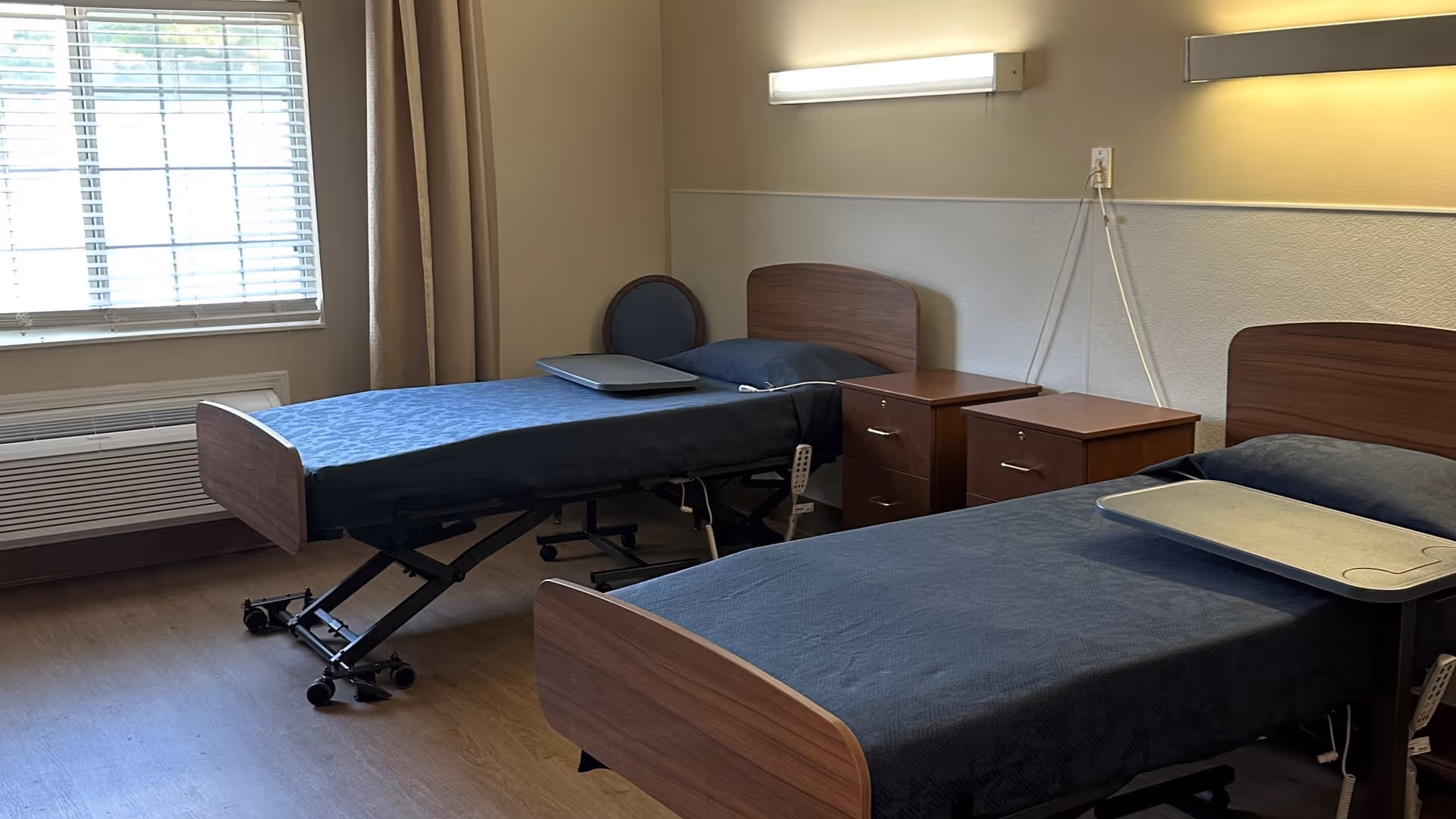 Two beds with blue blankets and nightstands in a shared senior living patient room.