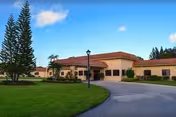 Single-story beige senior living facility with red-tiled roof, driveway, lawn, and palm trees.