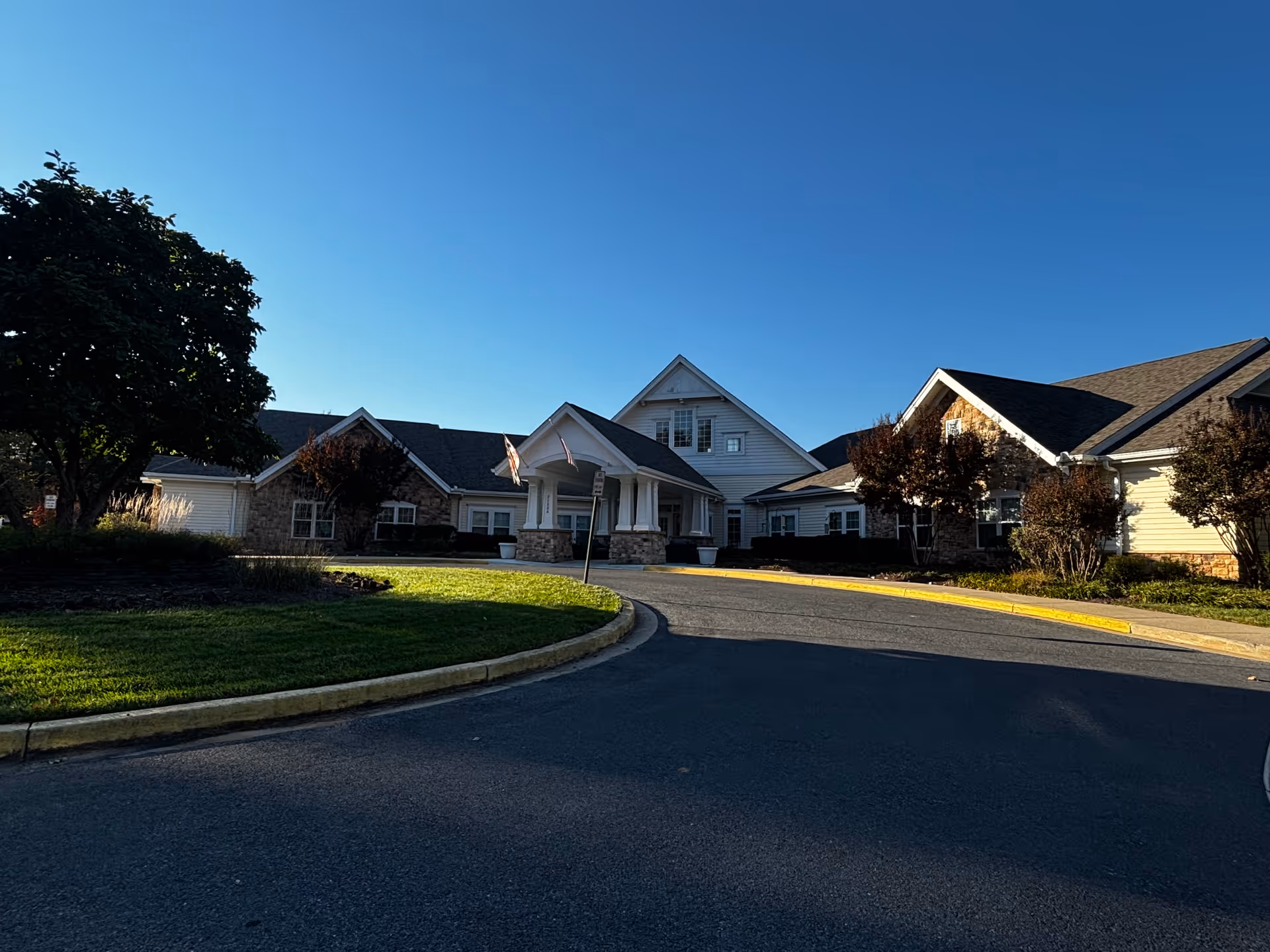 Exterior view of Somerford House Frederick, a senior living facility, showing a large building with a covered entrance, stone and siding facade, surrounded by trees and landscaping under a clear blue sky.