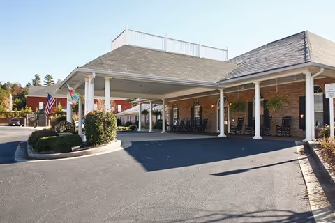 Brick senior living entrance with a covered driveway, rocking chairs, planters, and American flag.