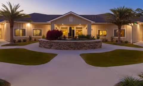 Front entrance of a single-story care facility at dusk with a circular stone planter, palm trees, and warm exterior lighting.