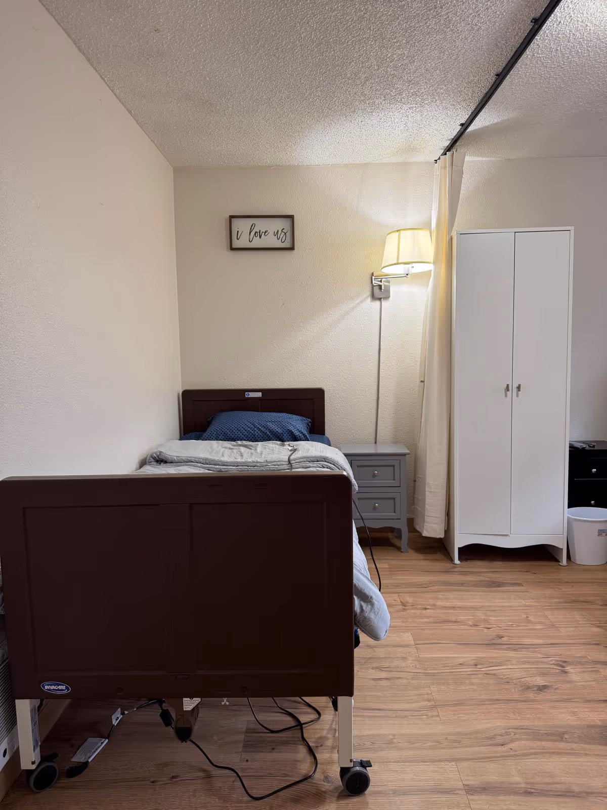 A small bedroom in a senior living facility with a bed, wardrobe, and bedside lamp.