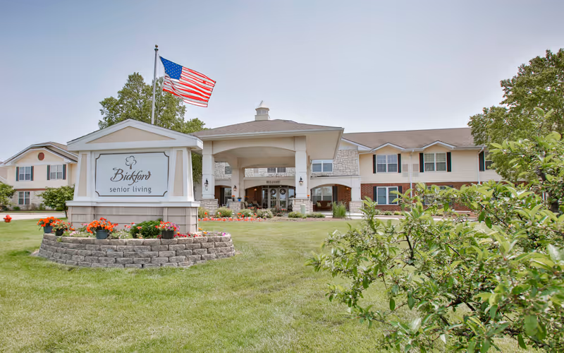 Front entrance of Bickford senior living with a sign, American flag, and landscaped lawn.