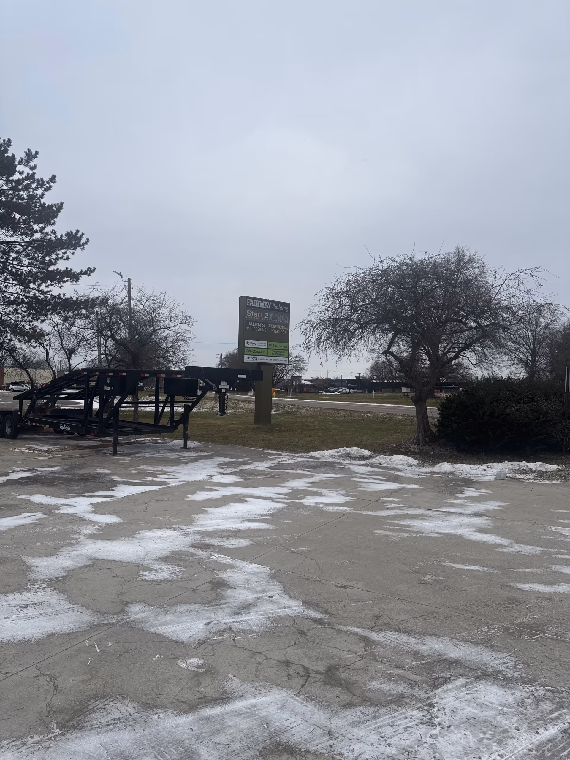 Overcast outdoor parking lot with a black trailer, leafless trees, and a sign.