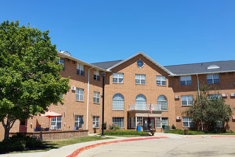 Front exterior of a three-story brick senior living building with arched windows, an entrance canopy, and trees under a clear blue sky.