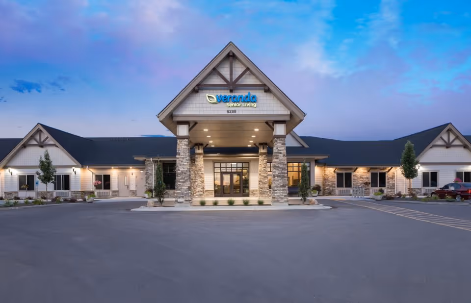 Front exterior of a single-story senior living building with a central covered entrance and illuminated windows under a twilight sky.