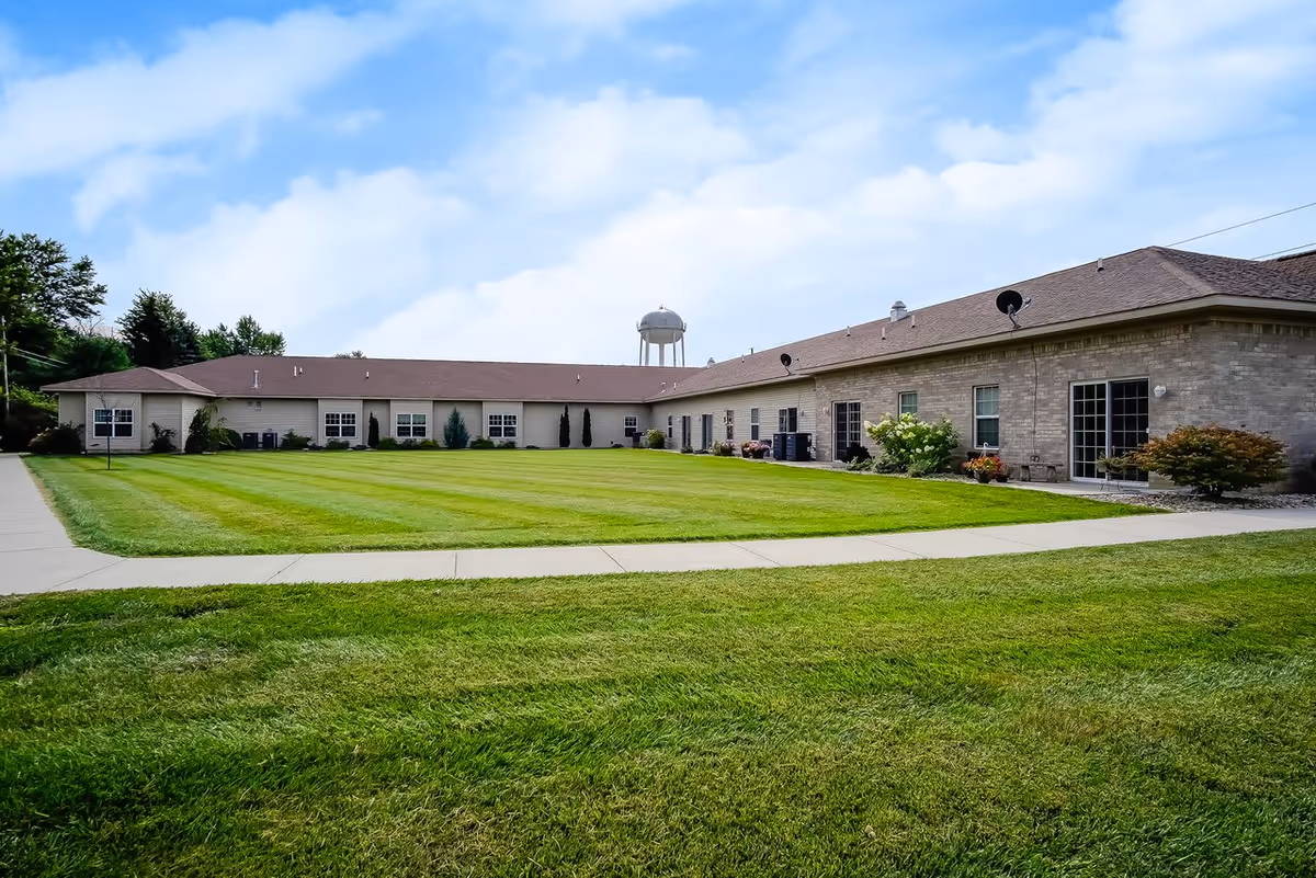 Exterior view of a single-story assisted living facility building with beige brick walls and a brown roof, surrounded by a well-maintained green lawn and a concrete walkway. A water tower is visible in the background under a partly cloudy blue sky.