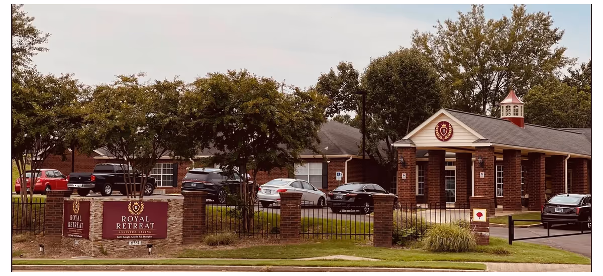 Front exterior of the Royal Retreat Assisted Living brick building with a driveway, parked cars, trees, and a sign in the foreground.